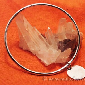 May include: A silver bangle bracelet with a round charm. The bracelet is on a red fabric background with a cluster of clear quartz crystals.