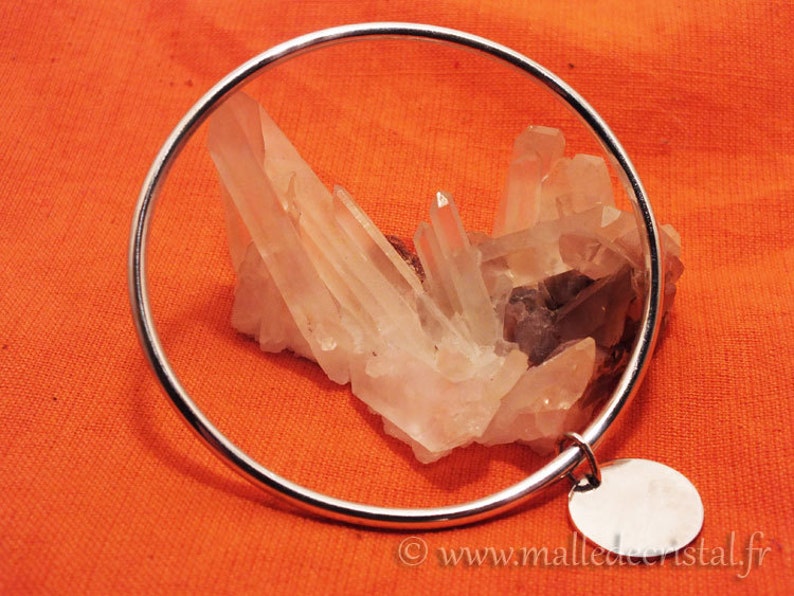 May include: A silver bangle bracelet with a round charm. The bracelet is on a red fabric background with a cluster of clear quartz crystals.