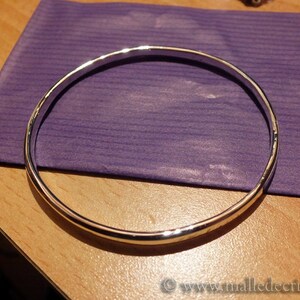 May include: A silver bangle bracelet with a hammered finish. The bracelet is lying on a purple surface.