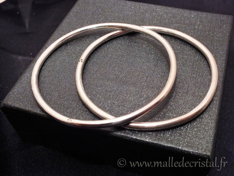 May include: Two silver bangle bracelets, one slightly larger than the other, are stacked on top of each other on a black surface.