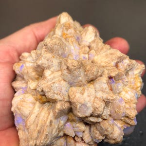 Puede incluir: Un espécimen de ópalo natural en bruto con una estructura cristalina en capas. La piedra presenta una mezcla de tonos beige y marrón, con destellos de púrpura y azul. El ópalo se sostiene en una mano, mostrando su tamaño y textura.