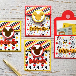 Puede incluir: Cuatro tarjeteros de regalo coloridos con un tema de Mickey Mouse. Los tarjeteros son rojos, amarillos y blancos, con un patrón repetido de cabezas de Mickey Mouse y estrellas. Cada tarjetero tiene un adorno de cabeza de Mickey Mouse y la palabra "Celebrate". Un bolígrafo dorado está en primer plano.