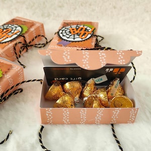 May include: Peach-coloured rectangular paper boxes with scalloped edges, decorated with a spiderweb and the word BOO. Each box contains gold-wrapped chocolate candies and a black gift card. The boxes are tied with black and gold twine.