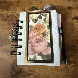 May include: A blue and white floral patterned journal with a gold trim and a spiral binding. The cover features a close-up of pink and yellow roses.