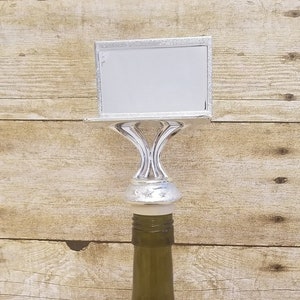May include: Silver bottle stopper with a blank rectangular sign on top. The stopper is on a green glass wine bottle.