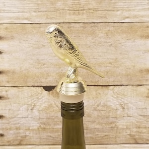 May include: Gold-colored metal bottle stopper shaped like a bird. The bird is perched on a round base with a cork stopper.