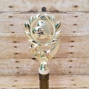 May include: Gold bottle stopper with a laurel wreath design and three stars. The center of the wreath features a football.