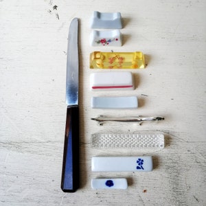 Puede incluir: Un juego de ocho palillos para comer sobre una superficie de madera blanca. Los palillos están hechos de diferentes materiales, incluyendo cerámica, plástico y metal. Los palillos están decorados con patrones florales y un diseño plateado.