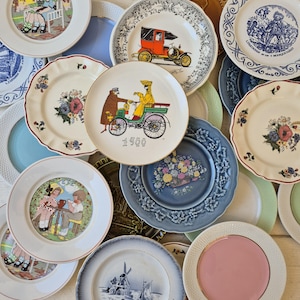 Op de afbeelding: Een verzameling decoratieve borden met diverse ontwerpen. Sommige borden tonen bloempatronen, terwijl andere scènes met vintage auto's en figuren afbeelden. De borden zijn in verschillende kleuren, waaronder wit, blauw en roze, en hebben verschillende randontwerpen.