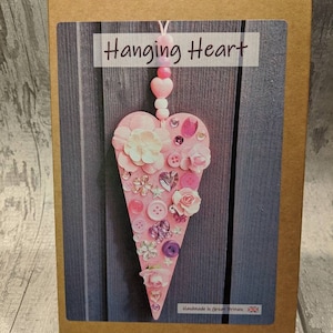 May include: A pink wooden heart decorated with a variety of pink, white and purple embellishments, including buttons, flowers and rhinestones. The heart is hanging from a string of pink beads. The box is labelled "Hanging Heart" and "Handmade in Great Britain".