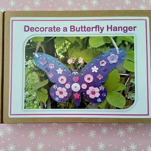 May include: A cardboard box with the text "Decorate a Butterfly Hanger" on the front. The box contains a purple butterfly-shaped craft project with colourful embellishments.