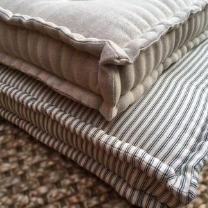 May include: Close-up of two stacked cushions. The top cushion is a solid light grey, with a tufted design and a ribbed edge. The bottom cushion has a white background with navy blue vertical stripes.