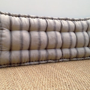 Puede incluir: Un cojín largo, rectangular, a rayas con un diseño capitoné. El cojín está hecho de una tela beige claro con rayas gris oscuro. El cojín está sobre una alfombra tejida.