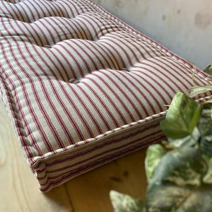 May include: A rectangular cushion with a cream-colored background and vertical red stripes. The cushion has a tufted design with buttons and a stitched border. The fabric appears to be cotton or linen, and the cushion is placed on a wooden surface.