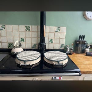 May include: A traditional black and cream-coloured stove with two covered burners. The stove has a black top and a cream-coloured base. The background includes a green wall, decorative tiles, and a wooden countertop.