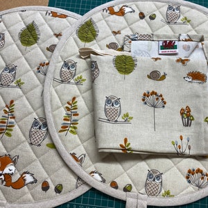 May include: Two round quilted pot holders with a brown and white owl design. A matching fabric tea towel with a brown and white owl design and the text "Made in Wales" is folded on top of the pot holders.