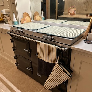 May include: A dark brown kitchen range with a stainless steel top, with two striped oven mitts and a striped towel. The range has multiple doors and a stainless steel handle. Two rectangular mats sit on the range top.