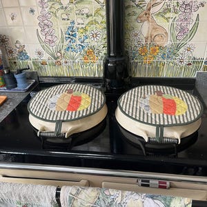 May include: Two round, cream-coloured stove top covers featuring a quilted fabric top with a pumpkin design in shades of orange, yellow, and grey. The covers have a dark green and white striped border and a central handle with a metal spring detail.