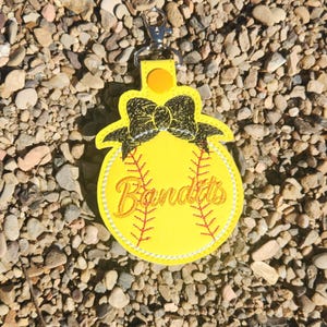 Puede incluir: Llavero de softball amarillo con un lazo negro y la palabra "Bandits" bordada.