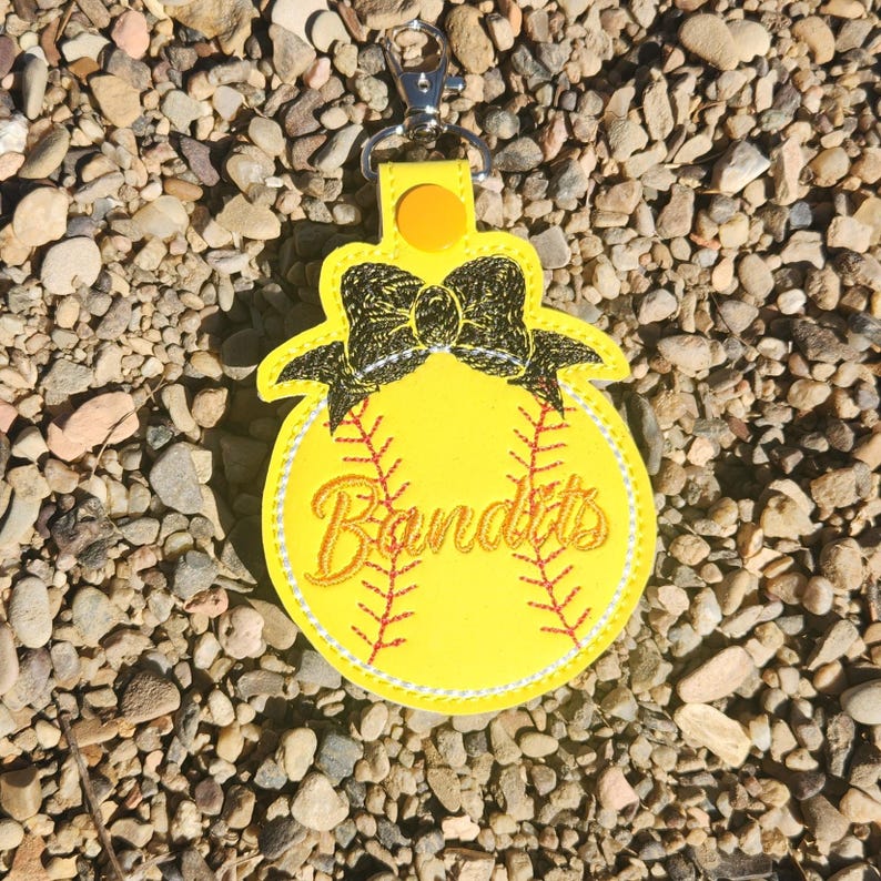 Puede incluir: Llavero de softball amarillo con un lazo negro y la palabra "Bandits" bordada.