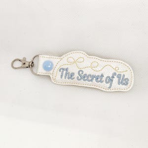 May include: White leather keychain with a blue button and gold embroidery. The text "The Secret of Us" is embroidered in blue thread.