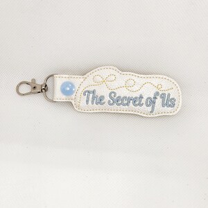May include: White leather keychain with a blue button and gold stitching. The keychain has the text "The Secret of Us" embroidered on it.