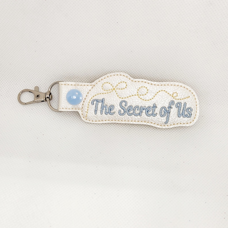 May include: White leather keychain with a blue button and gold stitching. The keychain has the text "The Secret of Us" embroidered on it.