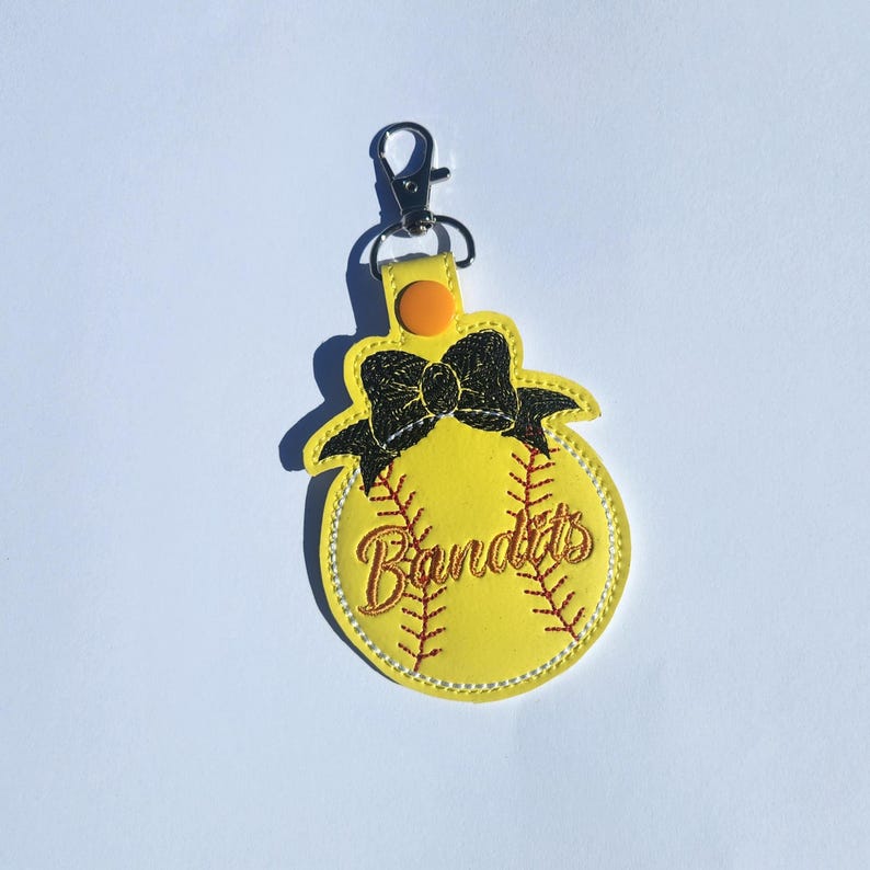 Puede incluir: Llavero de softball amarillo con un lazo negro y la palabra "Bandits" bordada en rojo.