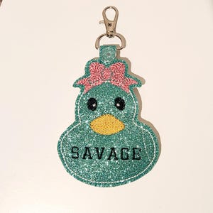 Pu&ograve; includere: Un'anatra di glitter blu turchese con un fiocco rosa e occhi neri. L'anatra ha un becco giallo e la parola "SAVAGE" &egrave; scritta in lettere nere sul davanti.