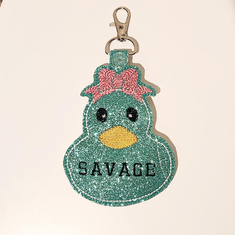 Pu&ograve; includere: Un'anatra di glitter blu turchese con un fiocco rosa e occhi neri. L'anatra ha un becco giallo e la parola "SAVAGE" &egrave; scritta in lettere nere sul davanti.