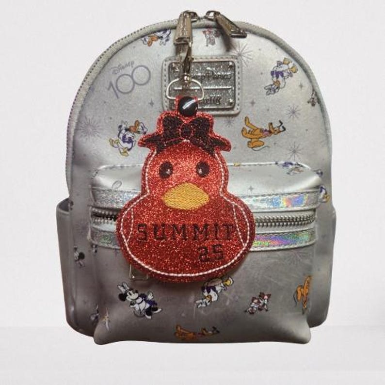 Pu&ograve; includere: Uno zaino grigio con un ciondolo a forma di anatra rosso glitterato. Lo zaino ha un logo Disney 100 bianco e diversi personaggi Disney stampati su di esso. Il ciondolo a forma di anatra ha il testo "Summit 25" su di esso.