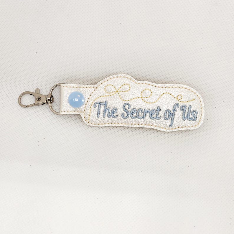 May include: White leather keychain with a blue button and gold stitching. The keychain has the text "The Secret of Us" embroidered on it.