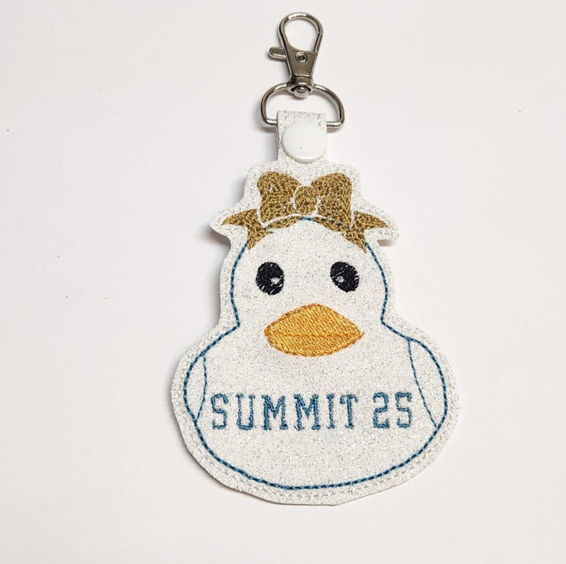 Pu&ograve; includere: Portachiavi a forma di papera di gomma bianca con un fiocco dorato, occhi neri e un becco arancione. La papera ha un ricamo blu attorno ai bordi e il testo "SUMMIT 25" sul davanti.