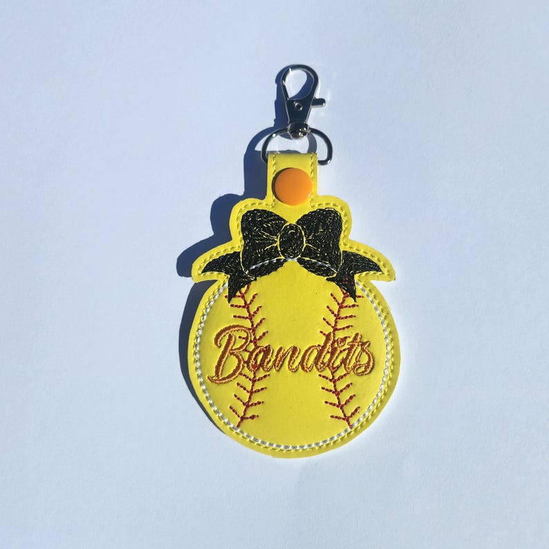 Puede incluir: Llavero de softball amarillo con un lazo negro y la palabra "Bandits" bordada en rojo. El llavero tiene un clip plateado.