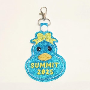 May include: A blue glitter duck-shaped keychain with a yellow bow and beak. The keychain has the text "SUMMIT 2025" embroidered in yellow. It includes a silver-colored metal clasp for attachment.