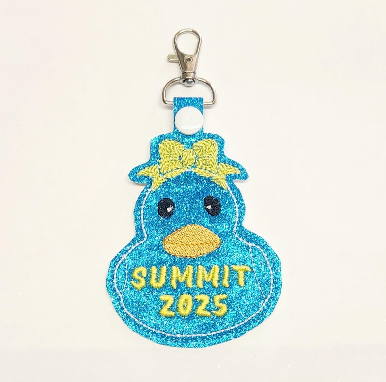 Pu&ograve; includere: Un portachiavi a forma di anatra blu glitterata con un fiocco giallo e il testo "SUMMIT 2025".