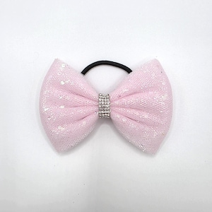 May include: A light pink hair tie featuring a bow made of glittery, mesh-like fabric. The centre of the bow is cinched with a band of small, clear, sparkling rhinestones. The elastic band attached to the back of the bow is solid black.