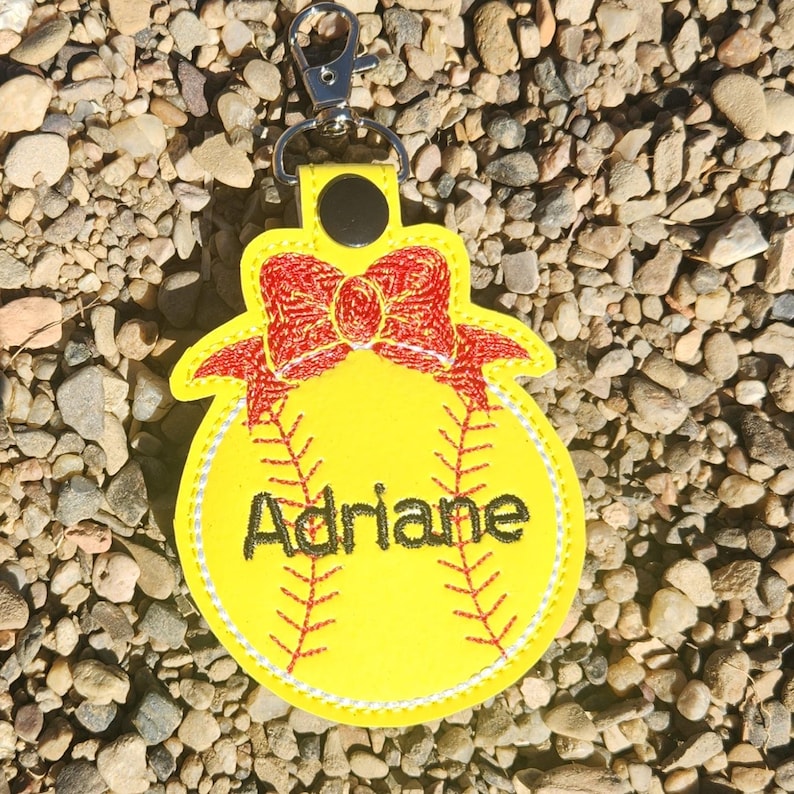 Puede incluir: Llavero de softball amarillo con un lazo rojo y el nombre Adriane bordado.