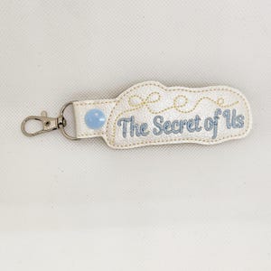 May include: White leather keychain with a blue button and the text "The Secret of Us" embroidered in blue thread.