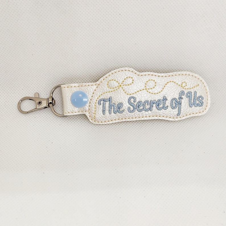 May include: White leather keychain with a blue button and the text "The Secret of Us" embroidered in blue thread.