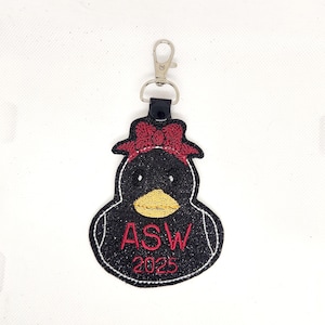 Pu&ograve; includere: Portachiavi a forma di papera di gomma nera con un fiocco rosso, glitter e il testo "ASW 2025" ricamato in rosso.