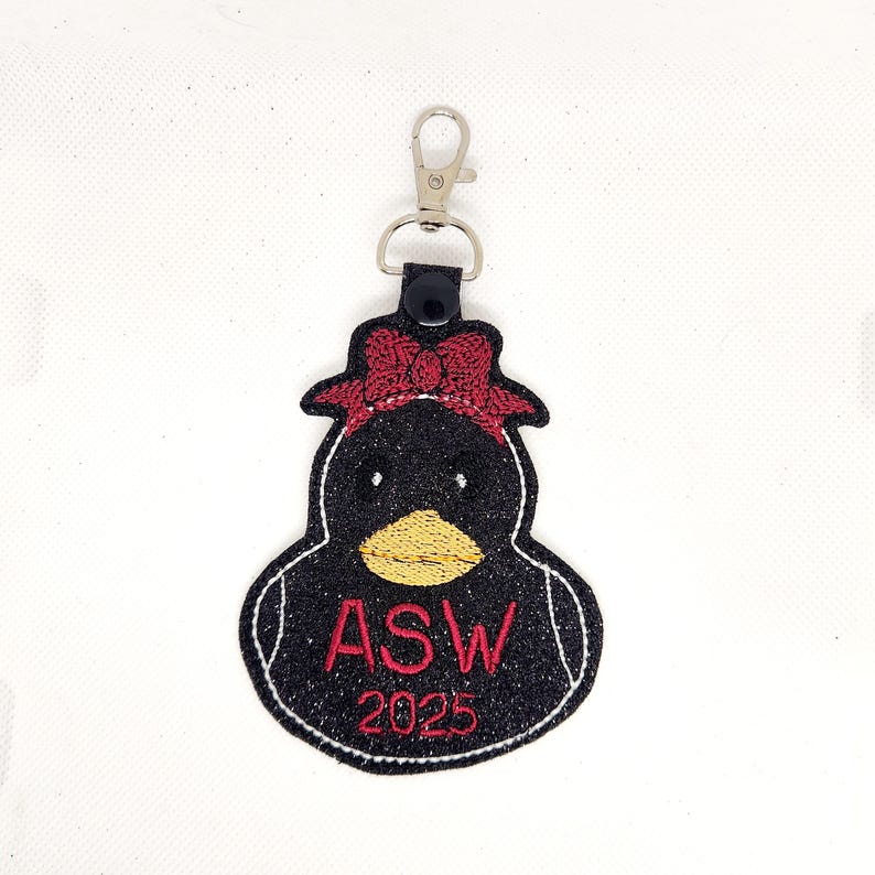 Pu&ograve; includere: Portachiavi a forma di papera di gomma nera con un fiocco rosso, glitter e il testo "ASW 2025" ricamato in rosso.