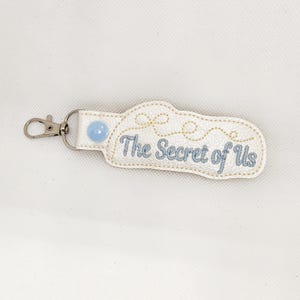 May include: White leather keychain with a blue button and gold embroidery that reads "The Secret of Us".