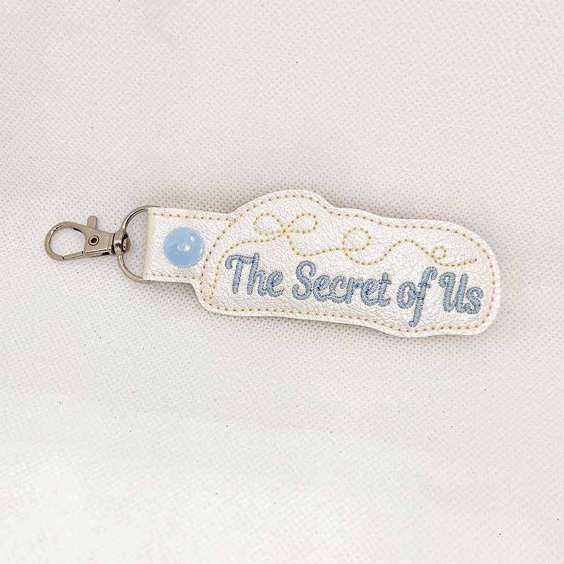 May include: White leather keychain with a blue button and gold embroidery that reads "The Secret of Us".