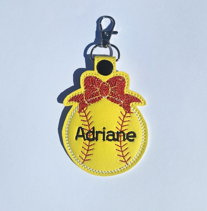 Puede incluir: Llavero de softball amarillo con un lazo rojo y el nombre Adriane bordado.