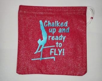 May include: Red drawstring bag with a white glitter finish. The bag features a blue silhouette of a gymnast on uneven bars with the text "Chalked up and ready to FLY!"