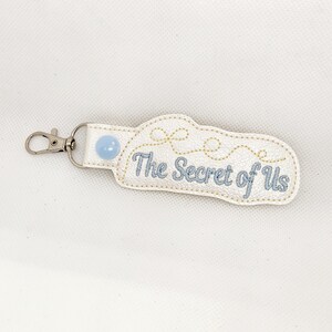 May include: White leather keychain with a blue button and gold embroidery. The text "The Secret of Us" is embroidered in blue thread.