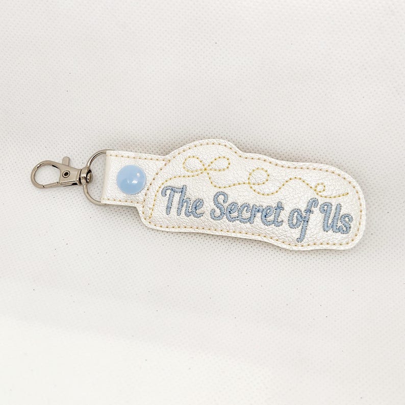 May include: White leather keychain with a blue button and gold embroidery. The text "The Secret of Us" is embroidered in blue thread.