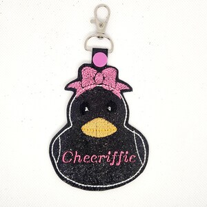 Pu&ograve; includere: Portachiavi pinguino nero e rosa con un fiocco rosa e la parola "Cheeriffic" ricamata sul davanti.