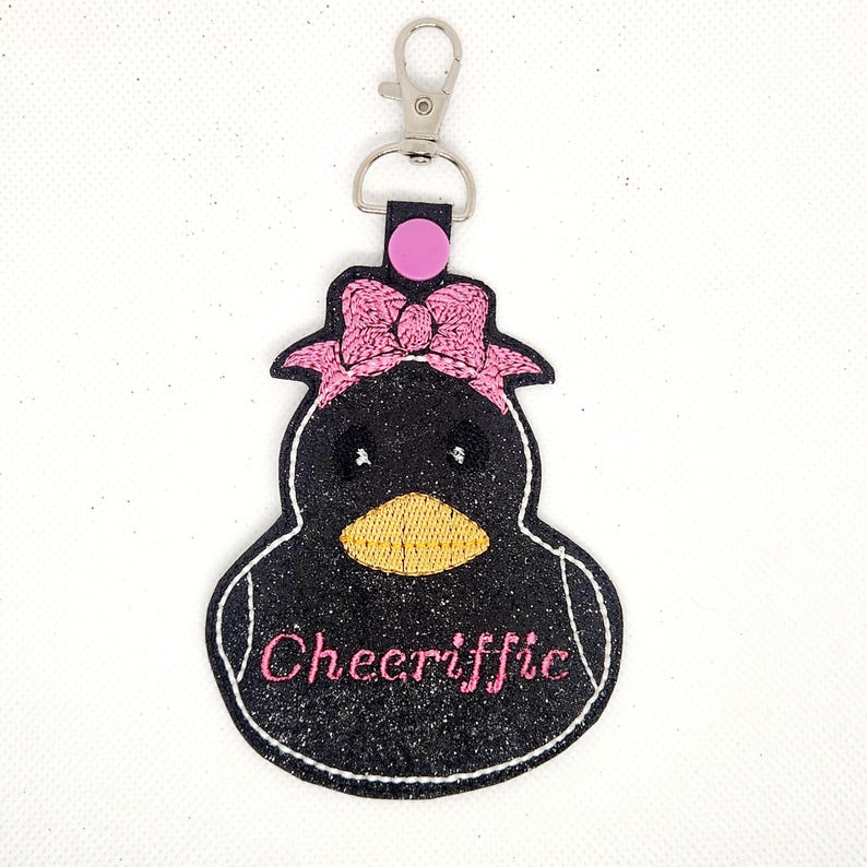 Pu&ograve; includere: Portachiavi pinguino nero e rosa con un fiocco rosa e la parola "Cheeriffic" ricamata sul davanti.